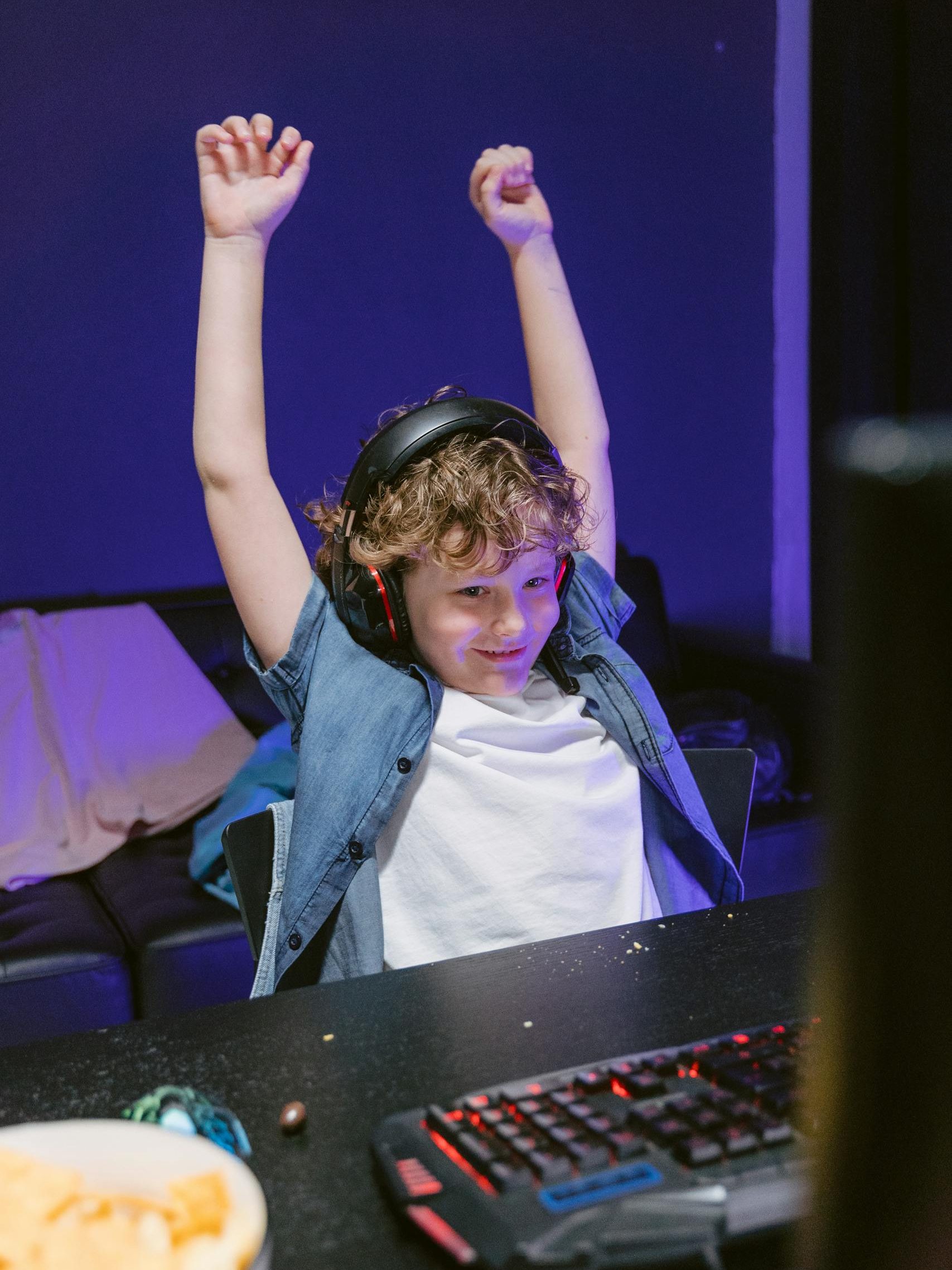 Child wearing headphones celebrates gaming victory with raised hands indoors.