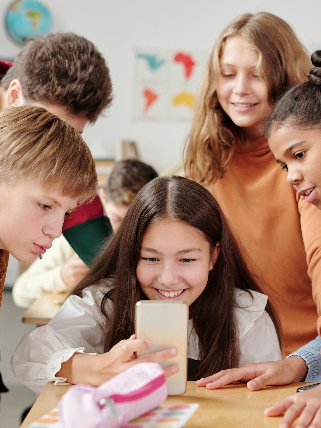 Group of teenagers in a classroom setting happily gathered around a smartphone.